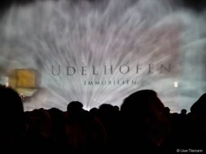 Menschen vor Wasserprojektion mit dem Schriftzug Udelhofen Immobilien.